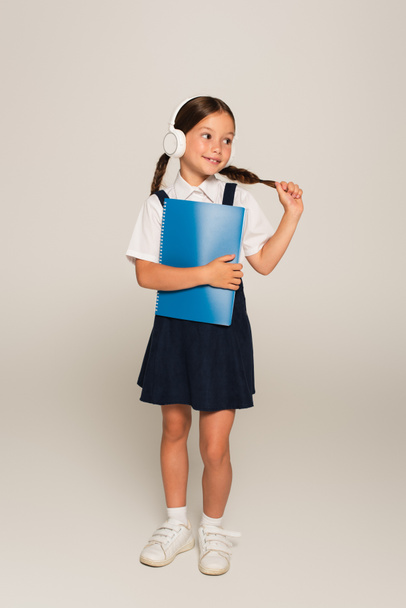 positive schoolgirl in headphones touching pigtail while holding copy book on grey - Foto, Bild