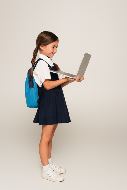 vista lateral de la muchacha en uniforme escolar usando el ordenador portátil en gris - Foto, imagen