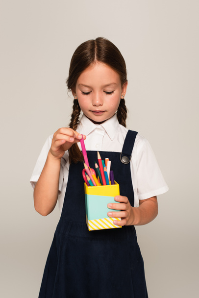 girl in school uniform taking felt pen from pen holder isolated on grey - Foto, Imagen