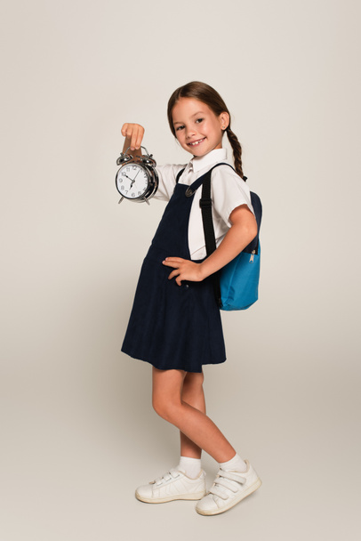 cheerful schoolgirl with blue backpack showing large alarm clock while standing with hand on hip on grey - Photo, Image