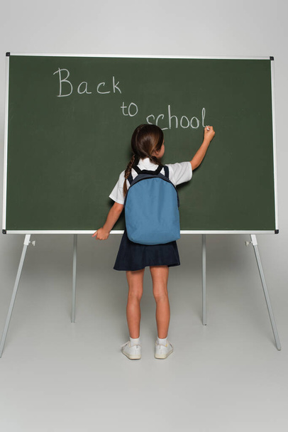 back view of schoolgirl with blue backpack writing back to school lettering on chalkboard on grey - Photo, Image