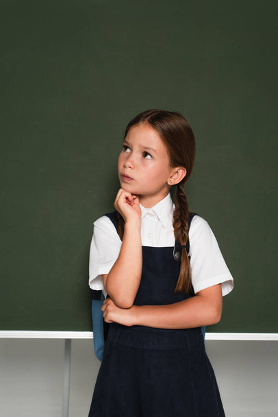 thoughtful schoolkid looking away and holding hand near face while standing at chalkboard on grey - Foto, immagini