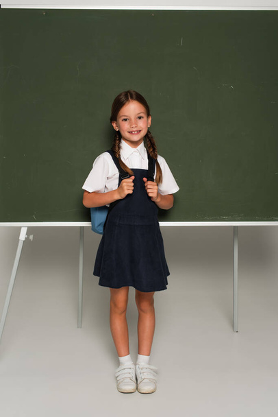 full length view of schoolchild with blue backpack smiling at camera near chalkboard on grey - Fotoğraf, Görsel