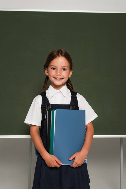 smiling schoolkid standing near chalkboard with notebooks on grey - Photo, image