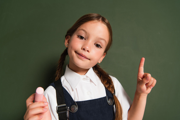 smiling schoolkid holding chalk and pointing at chalkboard while looking at camera - Foto, imagen