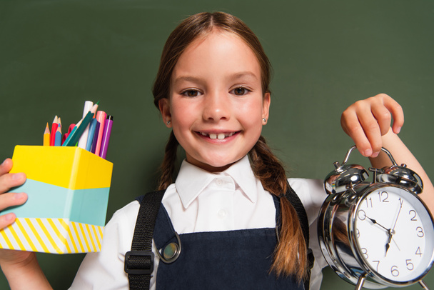 joyful schoolgirl showing pen holder and alarm clock near chalkboard - Photo, Image