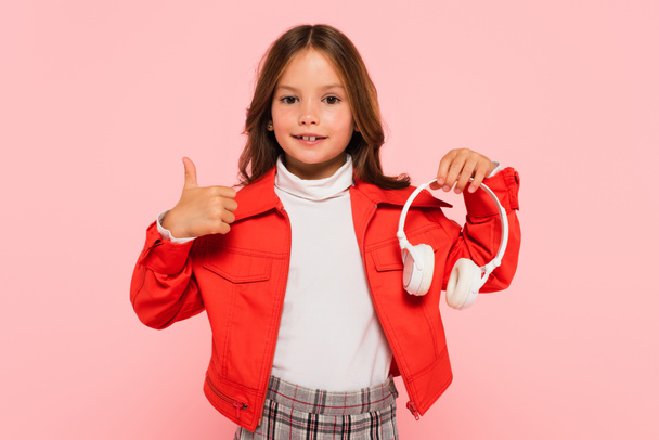 menina sorridente em jaqueta laranja elegante segurando fones de ouvido e mostrando polegar para cima isolado em rosa - Foto, Imagem