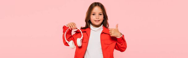stylish girl holding headphones and showing thumb up isolated on pink, banner - Foto, immagini