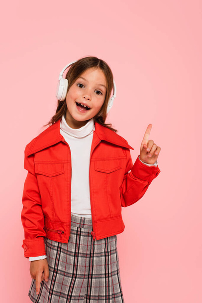 cheerful girl in orange jacket and headphones pointing with finger isolated on pink - Foto, afbeelding