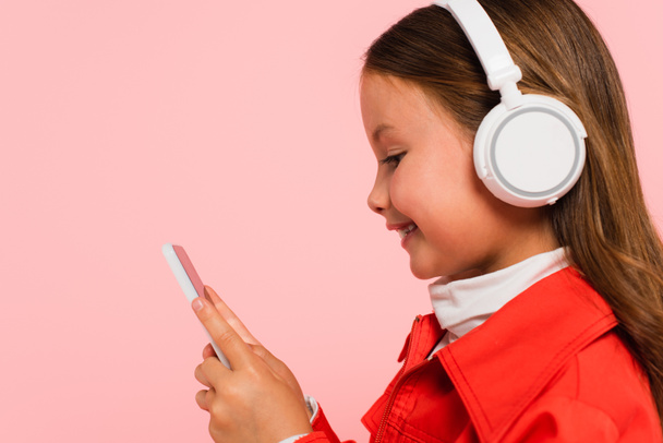 side view of happy child in headphones using smartphone isolated on pink - Foto, imagen
