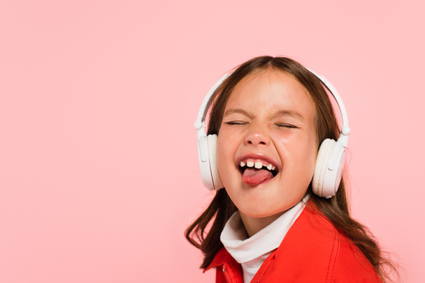 girl in headphones sticking out tongue with closed eyes isolated on pink - Photo, Image