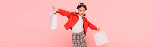 stylová dívka ve sluchátkách drží nákupní tašky izolované na růžové, banner - Fotografie, Obrázek