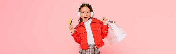innoissaan tyttö kuulokkeet ja tyylikäs asu poseeraa luottokortilla ja ostoskassit eristetty vaaleanpunainen, banneri - Valokuva, kuva