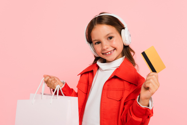 クレジットカードを持っているヘッドフォンの陽気な女の子とピンクに隔離されたショッピングバッグ - 写真・画像