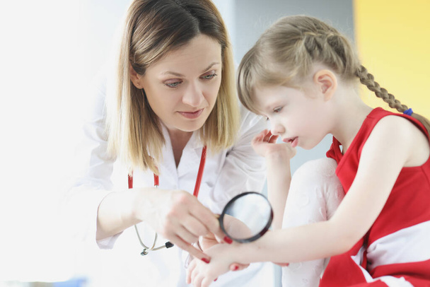 Доктор вивчає шкіру на руці маленької дівчинки, використовуючи збільшувальне скло в клініці
 - Фото, зображення