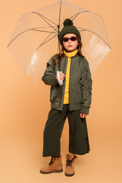 full length view of girl in bomber jacket and suede boots under transparent umbrella on beige - Фото, изображение