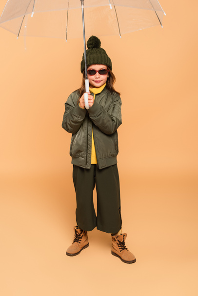 full length view of kid in sunglasses and stylish clothes under transparent umbrella on beige - 写真・画像
