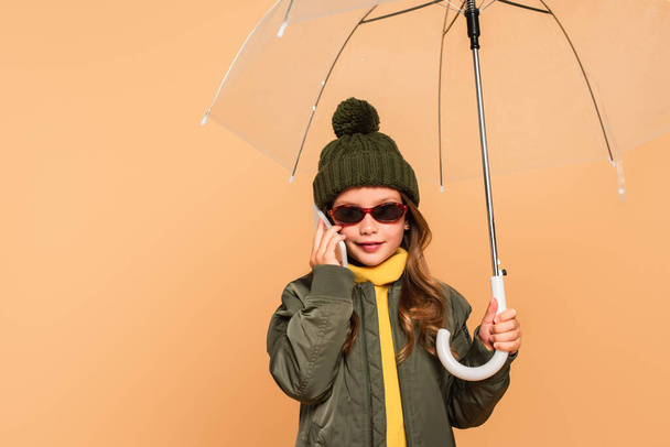 bambino elegante in occhiali da sole che parla su smartphone sotto l'ombrello isolato sul beige - Foto, immagini