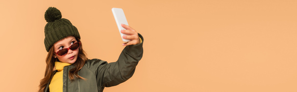 Trendgirl mit Strickmütze und Sonnenbrille macht Selfie isoliert auf beige, Banner - Foto, Bild