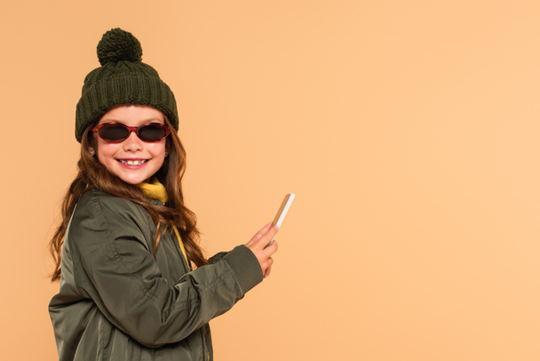 cheerful kid in sunglasses, jacket and knitted hat holding mobile phone isolated on beige - Photo, Image