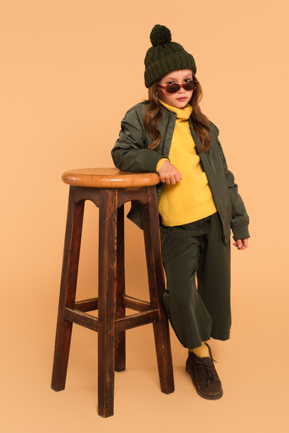 child in yellow turtleneck and bomber jacket posing near high stool on beige - Φωτογραφία, εικόνα