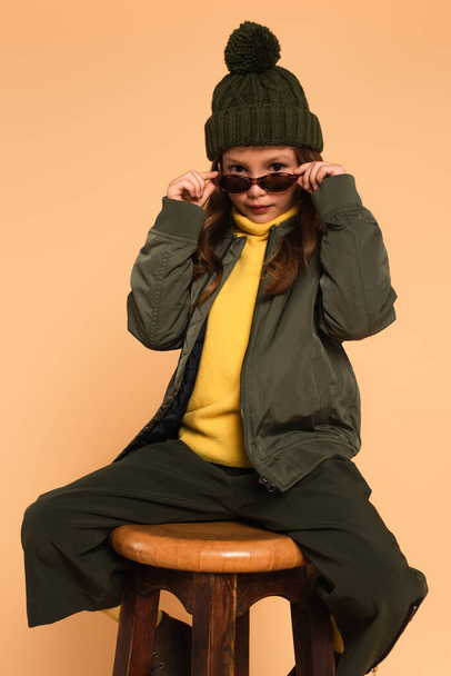 criança em roupa de outono na moda olhando para a câmera sobre óculos de sol elegantes isolados no bege - Foto, Imagem