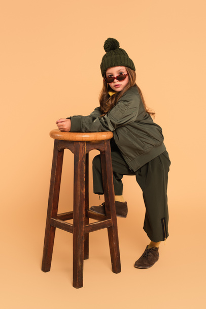 fashionable girl in autumn clothes looking at camera over sunglasses near high stool on beige - Foto, immagini