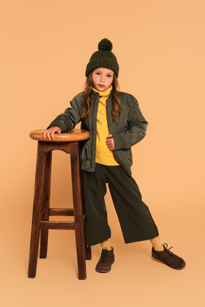 stylish child in autumn outfit looking at camera near high wooden stool on beige - Photo, Image