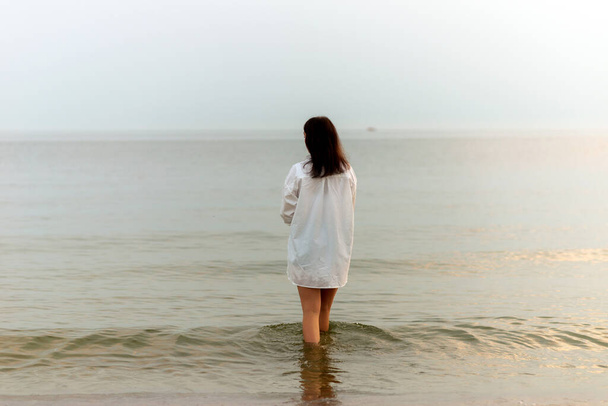mooie vrouw wandelen langs strand en zee zonsondergang achtergrond.Vrouw in jurk wandelen op oceaan.Eenzaamheid en verdriet concept.Lege plek voor emotionele, sentimentele, inspirerende tekst. Achteraanzicht. - Foto, afbeelding