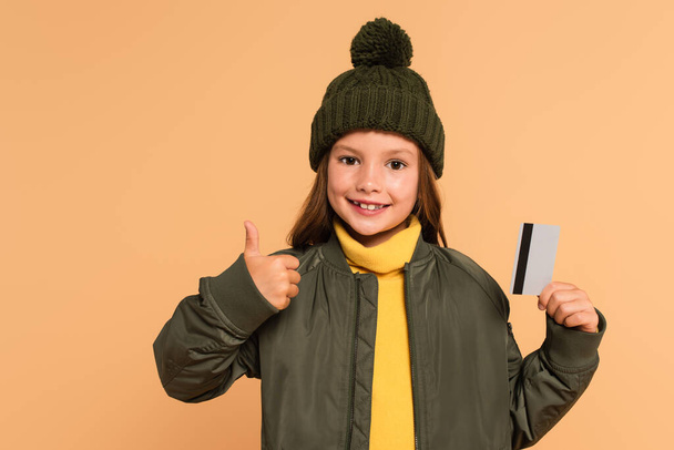 glückliches und stilvolles Kind zeigt den Daumen nach oben, während es die Kreditkarte isoliert auf beige hält - Foto, Bild