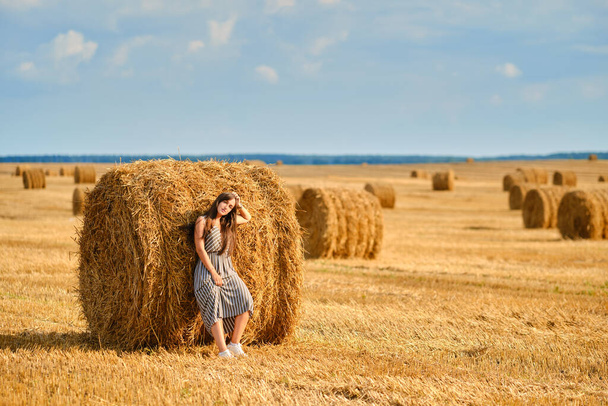 Happy young woman leaning to the round stack of straw on the field - Foto, Imagen