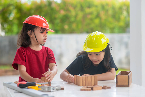 Asijské sourozenci dívky nosí strojírenské klobouky budova dům z dřevěné hračky. Na učení a rozvoj, malý architekte. - Fotografie, Obrázek