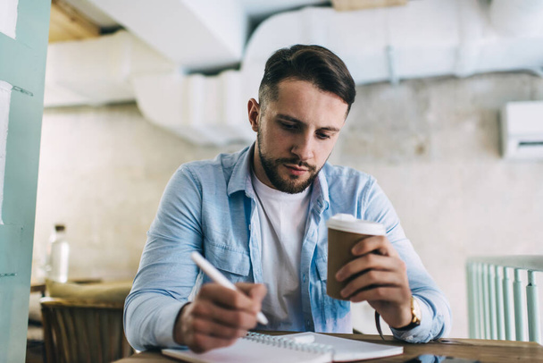 Serious bearded man in casual clothes sitting at table in modern cafe and making notes in copybook while drinking hot coffee - Fotó, kép