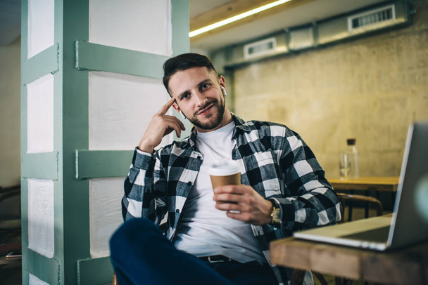 Smiling male hipster in checkered shirt sitting at table with takeaway coffee and laptop and listening to songs in true wireless earphones while chilling and looking at camera - Фото, зображення