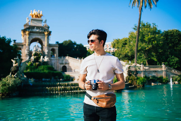 Stylish ethnic male traveler with belt bag standing on background of historic fountain in park during summer vacation in Barcelona and looking away - 写真・画像
