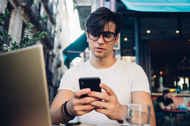 Concentrated charismatic man in glasses and bracelets sitting at cafe in Barcelona using smartphone and text messaging while working outside - Fotografie, Obrázek