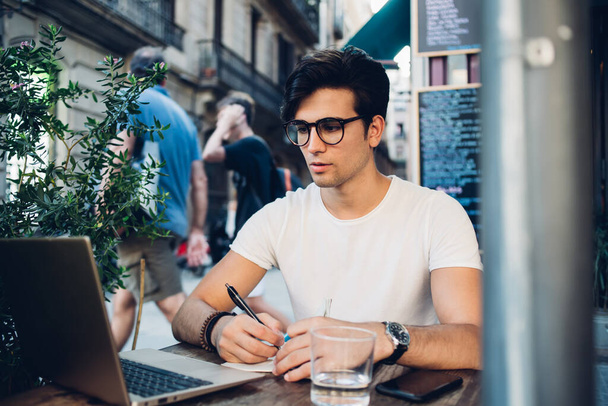 Serious caucasian man in eyewear for vision protection learning via online course making notes in notepad, pensive handsome male student watching training educational webinar on cafe terrace - Foto, afbeelding