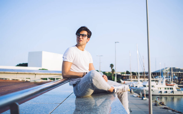 Handsome caucasian male in trendy wear resting outdoors on weekends looking at yachts and boats in bay, pensive 20s hipster guy traveler enjoying leisure and summer journey near marina in port - Фото, зображення