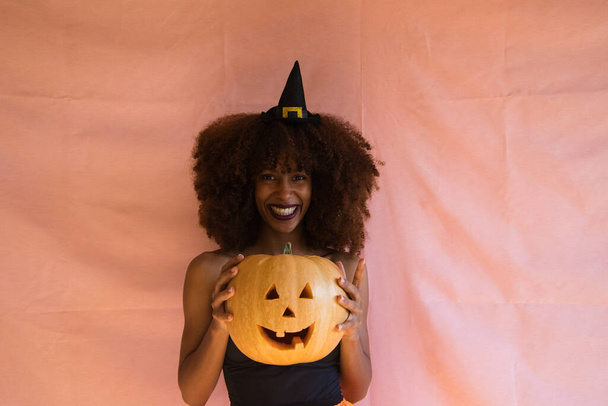 nuori afrikkalainen amerikkalainen nainen pukeutuneena noidaksi halloween-juhlia varten ja pitäen kurpitsaa käsissään. Hänellä on noidan hattu ja oranssi hame. Nainen hymyilee kameralle kurpitsan kanssa.. - Valokuva, kuva