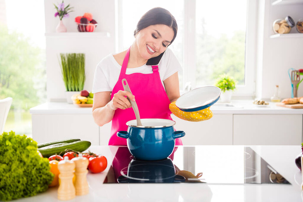Photo portrait woman in pink apron smiling talking on cellphone cooking soup - Photo, image
