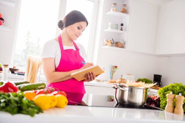 Photo portrait woman in pink apron writing notes in blocknote cooking pasta - Foto, Imagem
