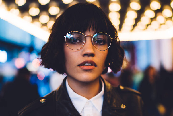 Close up portrait of female model dressed in trendy clothing have evening sightseeing around illuminated street in megalopolis, stylish hipster girl in glasses looking at camera during travel - Foto, Imagen