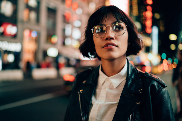 Caucasian hipster girl in classic spectacles for provide eyes protection thinking about metropolitan nightlife in New York, thoughtful female in fashionable outfit recreating during evening in city - Φωτογραφία, εικόνα
