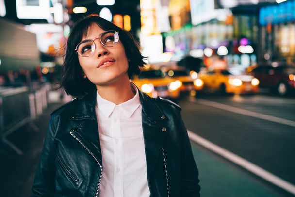 Charming brunette young woman in stylish spectacles looking up thinking at urban setting of night city,pensive hipster girl in leather jacket pondering about nightlife in metropolis standing at street - Фото, изображение