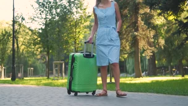 Молодая женщина идет по парку тротуар с зеленым чемоданом в летний день. Концепция путешествий и отдыха. - Кадры, видео