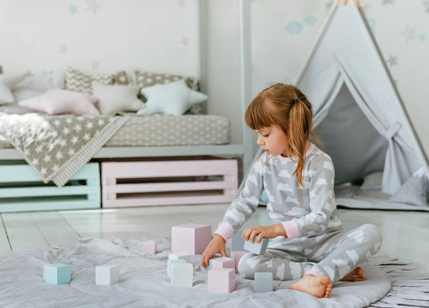 Beautiful little girl wearing pajamas playing with wooden toy blocks in the kids room. Natural eco toys concept. Selective focus. - Фото, изображение