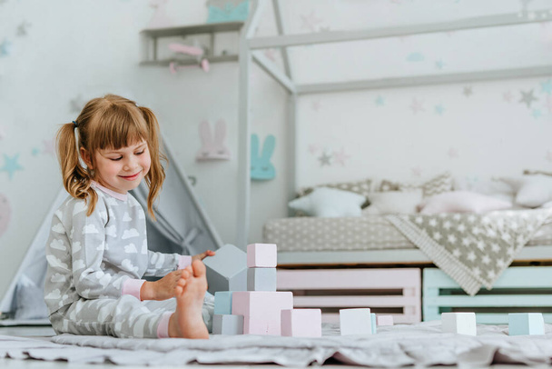 Mooi klein meisje met een pyjama spelend met houten speelgoed blokken in de kinderkamer. Natuurlijke eco speelgoed concept. Selectieve focus. - Foto, afbeelding