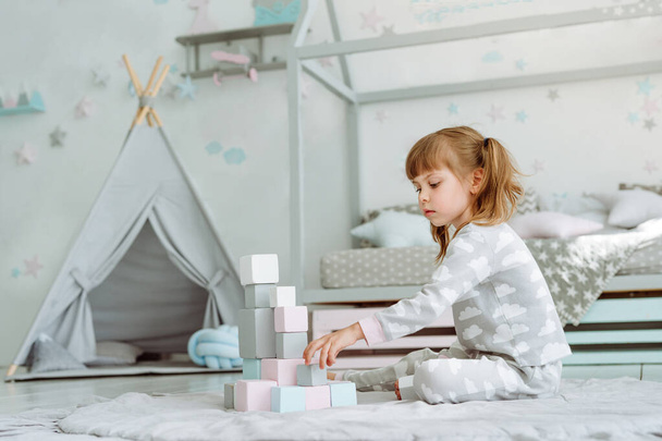 Piękna dziewczynka w piżamie bawiąca się drewnianymi klockami w pokoju dziecięcym. Koncepcja naturalnych zabawek ekologicznych. Skupienie selektywne. - Zdjęcie, obraz