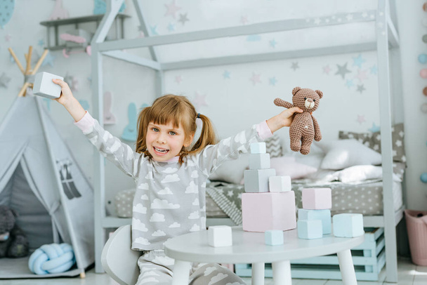 Mooi klein meisje met een pyjama spelend met houten speelgoed blokken en berenspeelgoed zittend aan tafel in de kinderkamer. Natuurlijke eco speelgoed concept. Selectieve focus. - Foto, afbeelding