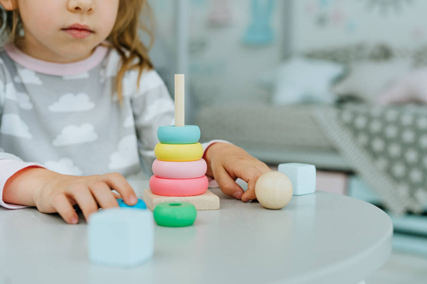 Close up of little toddler girl wearing pajamas playing with wooden toy pyramid sitting at the table in the kids room. Natural eco toys concept. Selective focus. - Фото, изображение
